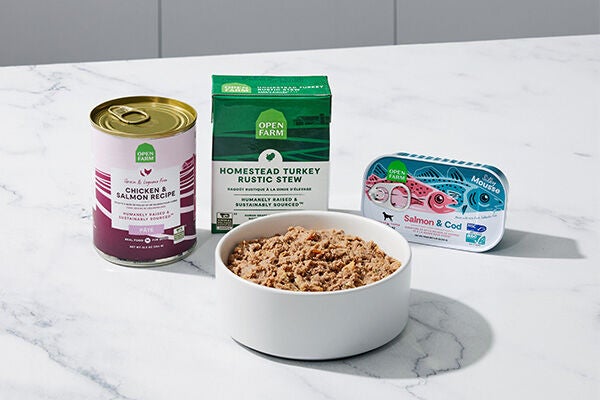 Three containers of wet open farms dog food, one in a pink can, one in a green box, and one in a tin with fish on it sitting behind a white bowl filled with wet dog food on a white marble counter